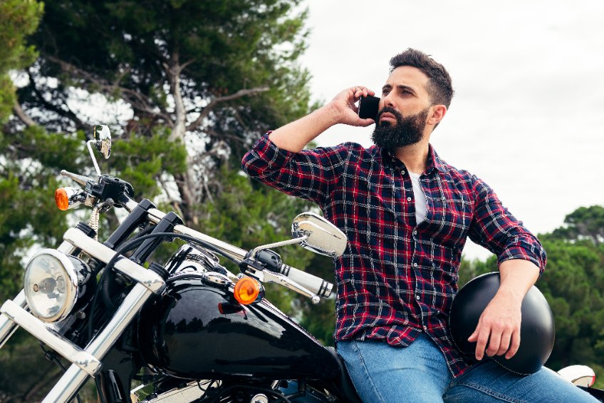 Motard au téléphone, assis de profil sur le siège de sa moto noire garée, avec son casque sous le bras